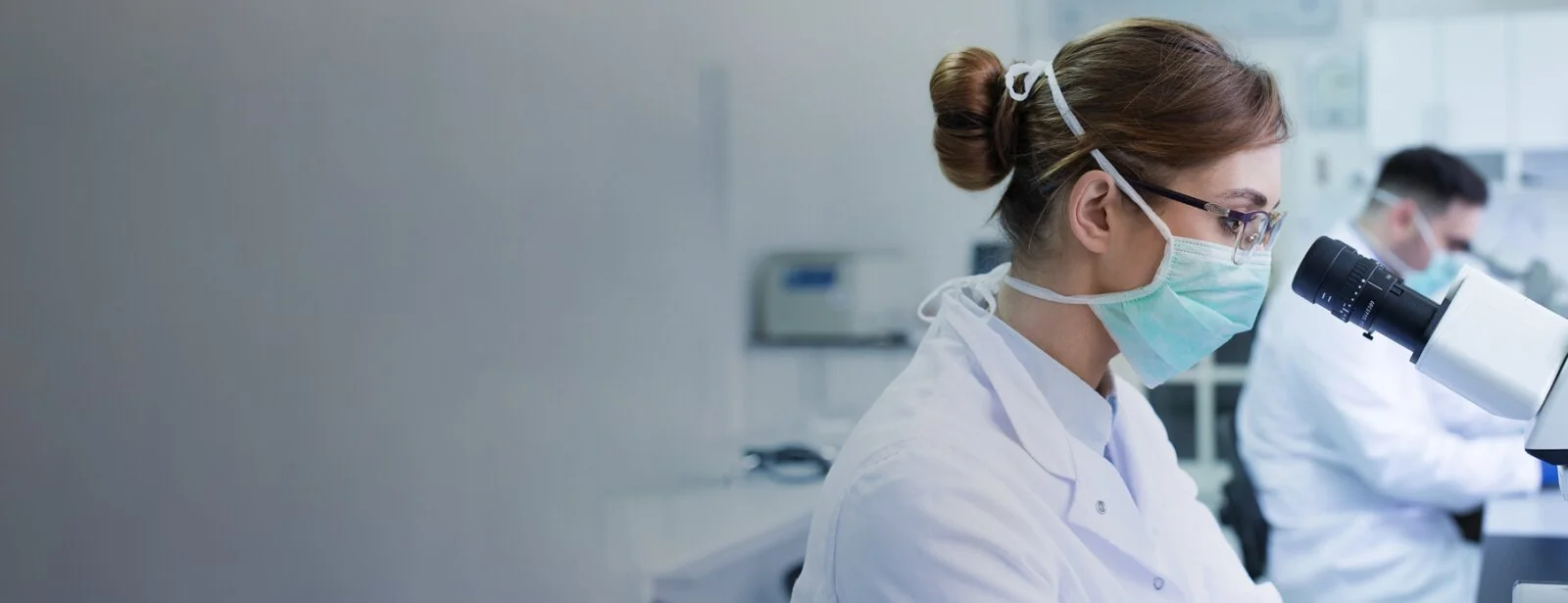 Focus on a doctor looking into a microscope. Another doctor is working in the background.